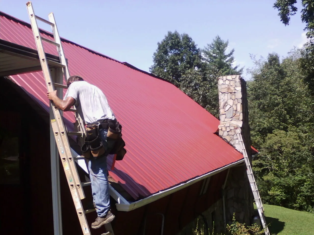 Roofer on a ladder beside a red metal roof and stone chimney — Soffit & Fascia Repair by Roofing Brothers in Fort Washington